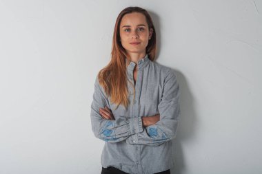 Açık gri elbiseli, mavi gözlü, sarı saçlı ve yüzünde bir gülümseme olan kendinden emin bir kadın kollarını kavuşturmuş beyaz duvarın yanında duran kameraya bakıyor. İnsanlar, gençlik, güzellik, yaşam tarzı konsepti 