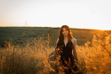 Gün batımı, yumuşak altın sarısı akşam renkleri, genç, güzel, duygusal esmer uzun siyah uzun elbiseli kız altın buğday tarlasında. Ruhun rahatlığı, neşe, doğa