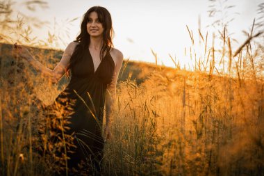 Gün batımı, yumuşak altın sarısı akşam renkleri, genç, güzel, duygusal esmer uzun siyah uzun elbiseli kız altın buğday tarlasında. Ruhun rahatlığı, neşe, doğa