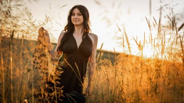 Gün batımı, yumuşak altın sarısı akşam renkleri, genç, güzel, duygusal esmer uzun siyah uzun elbiseli kız altın buğday tarlasında. Ruhun rahatlığı, neşe, doğa