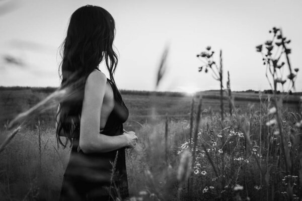 Sunset, on a wheat field a young, beautiful, emotional girl - a brunette with long hair turned her back and goes to the sun. Peace, joy, nature, black and white photo