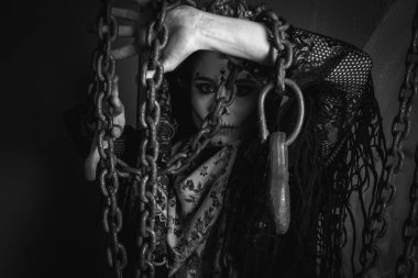 Mutlu Cadılar Bayramı konsepti. Korkunç bir kadın portresi, özel kıyafetli, profesyonel makyajlı, korkutucu görünmek için siyah elbiseli, gri arka plandaki boş bir alanda, siyah beyaz bir fotoğrafta.