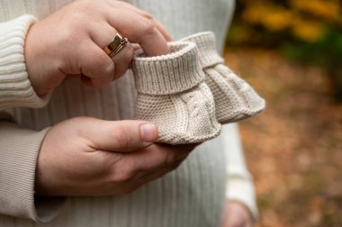 Doğurmak üzere olan anne yakın plan örgü elbisesiyle şişliğini sallıyor ve dışarıda sonbaharda örülmüş bebek paçalarını tutuyor.