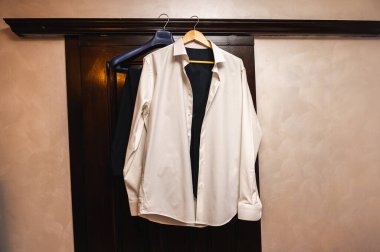 Ahşap bir kapı çerçevesinde asılı duran resmi beyaz gömlek ve siyah yelek. Düğünden ya da iş etkinliğinden önce damat hazırlama konsepti. Zarif erkek giyim geçmişi