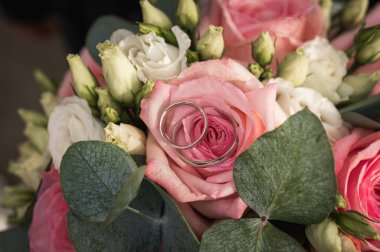 Beyaz çiçekli ve okaliptüs yapraklı bir gelin buketinde pembe gül yapraklarının üzerinde duran iki gümüş alyansın yakın çekimi. Aşk ve evliliğin romantik sembolü, seçici odaklanma