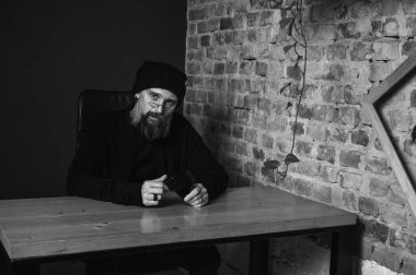 Ciddi sakallı, gözlüklü bir hippinin çatı katındaki ahşap bir masada oturan siyah beyaz portresi. Tuğla duvara bakan kendine güvenen bir serbest yazar.