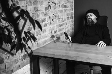 Ciddi sakallı, gözlüklü bir hippinin çatı katındaki ahşap bir masada oturan siyah beyaz portresi. Tuğla duvara bakan kendine güvenen bir serbest yazar.