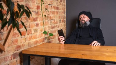 Gözlüklü sakallı hippi çatı katındaki ahşap masada otururken akıllı telefon kullanıyor. Kırmızı tuğlalı arka planda sıradan bir erkek telefonu. Uzak çalışma
