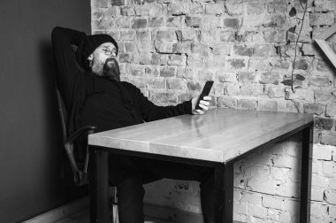 Ciddi sakallı, gözlüklü bir hippinin çatı katındaki ahşap bir masada oturan siyah beyaz portresi. Tuğla duvara bakan kendine güvenen bir serbest yazar.