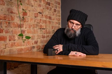 Ciddi sakallı, gözlüklü ve bereli bir hippinin portresi akıllı telefonuyla masada oturuyor. Çatı katında tuğla duvar arkasında yaratıcı bir serbest çalışan.