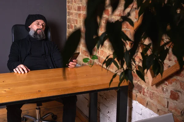 Gözlüklü sakallı hippi çatı katındaki ahşap masada otururken akıllı telefon kullanıyor. Kırmızı tuğlalı arka planda sıradan bir erkek telefonu. Uzak çalışma