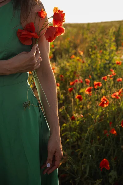 Yeşil elbiseli, kırmızı gelincik çiçekleri taşıyan bir kadının yakın çekimi. Yaz çayır arka planı ve gün batımı ışığı. Kadın güzelliği, şefkat ve doğa kavramı.