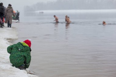Suda yıkanan insanların arka planında kış nehrinin kenarında oturan bir çocuk.
