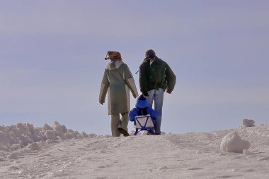 Bir adam, bir kadın ve bir çocuk kar üzerinde mavi gökyüzüne karşı duruyor.