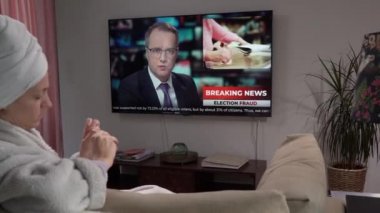 Genç bir kadın manikür yapıyor. Oturma odasında, elinde televizyonla oturuyor..