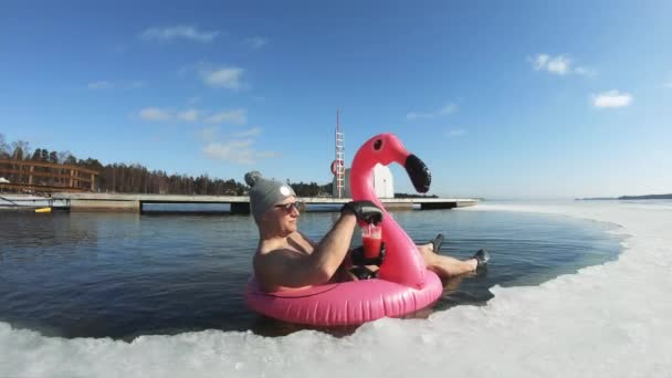 Um homem sonhando com umas férias flutuando em um flamingo inflável em um buraco de gelo — Vídeo de Stock