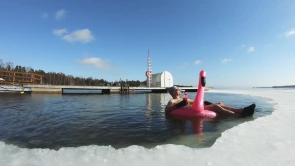 Um homem sonhando com umas férias flutuando em um flamingo inflável em um buraco de gelo — Vídeo de Stock