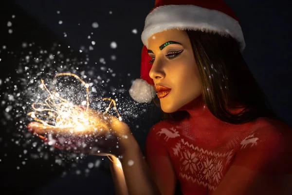 Krásná brunetka s uměleckým make-upem, v klobouku Santa Claus, stojí s úsměvem na modrém pozadí ve studiu — Stock fotografie