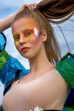 Gökyüzüne karşı mavi yağmurluk giymiş, uzun saçlı güzel bir kadının yaratıcı, modaya uygun bir portresi. Profesyonel makyaj..