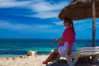 Bir kız, Akdeniz kıyısındaki bir şemsiyenin altında bir şezlongda oturur ve huş ağacı suyuyla denize bakar..