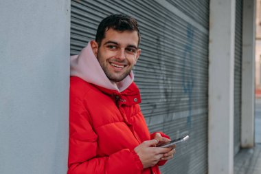 Duvara yaslanmış gülümseyen cep telefonlu genç adam.