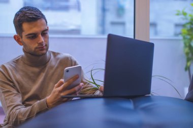 Evde dizüstü bilgisayarı ve akıllı telefonuyla çalışan çekici bir genç.