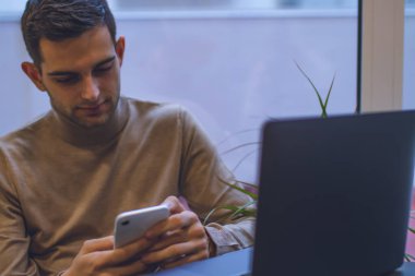 Evde dizüstü bilgisayarı ve akıllı telefonuyla çalışan çekici bir genç.