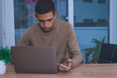 Evde dizüstü bilgisayarı ve akıllı telefonuyla çalışan çekici bir genç.