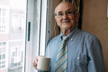 Evde elinde bir fincan kahveyle pencereden bakan son sınıf öğrencisi.