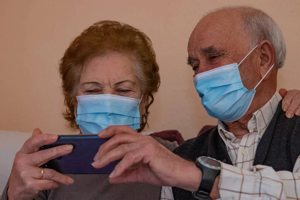 older couple with mobile phone talking on video conference with masks