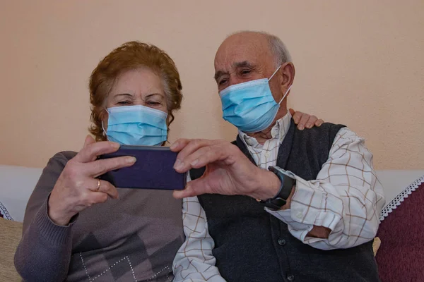 older couple with mobile phone talking on video conference with masks