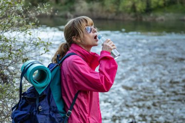 Sırt çantalı kadın içki içiyor, spor yapıyor ya da yürüyüş yapıyor.