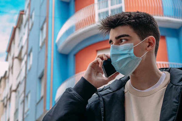 Young male teenager with mobile phone and mask on the street with buildings