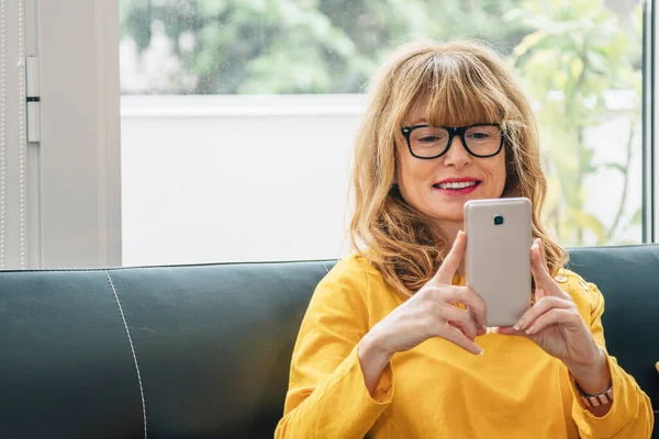 Evdeki Kadın Koltukta Oturan Cep Telefonuna Bakıp Gülümsüyor — Stok fotoğraf