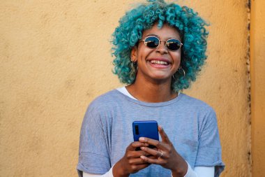 Renkli duvarda cep telefonu olan mavi saçlı Afro-Amerikalı bir kız.