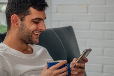 Elinde kahve olan genç adam cep telefonuna bakıyor.