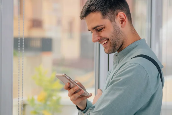 Ung Man Tittar Mobilen Bredvid Fönstret Leende — Stockfoto