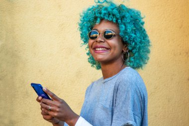Sarı duvar arkasında cep telefonu ve güneş gözlüğü olan Afro-Amerikan Latin kız.