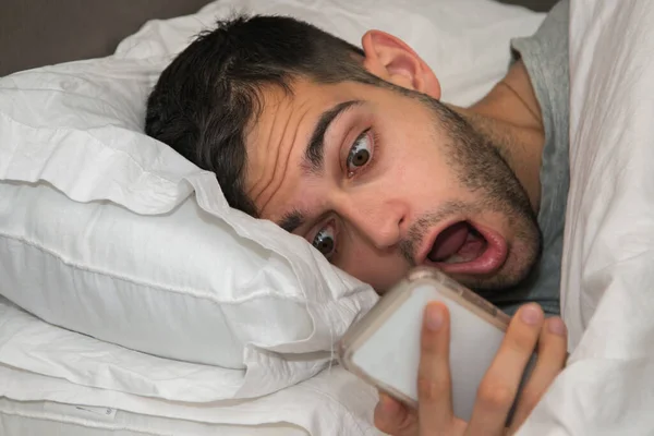 young man in bed staring at the phone, scared or surprised