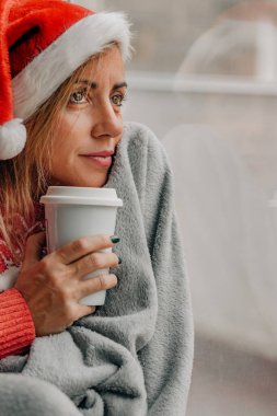 Noel 'de pencerede battaniye ve kupayla Noel Baba şapkası takan bir kadın.