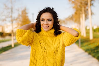 Parlak sarı bir süveter içinde inanılmaz güzel bir gülümseme ile beyaz dişli kıvırcık saçlı esmer ellerini başının arkasına attı ve sonbaharda parkın arka sokağında durup gülümsedi.