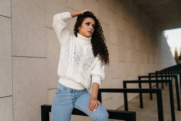 Pięknie pozuje fotograf brunetka w dżinsach i długi sweter, podnosząc rękę kręconymi włosami — Zdjęcie stockowe