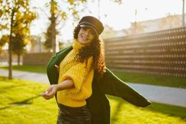 Şapkalı ve ceketli neşeli bir kız parkta dönüyor ve kameraya gülüyor. Sonbahar