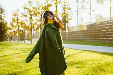 Yeşil ceketli güzel bir kız parkta yürür, elini şapkaya koyar ve kameraya döner..