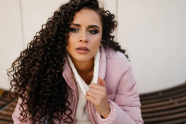 Beyaz kazaklı, düşüncelere dalmış kıvırcık bir kadın dışarıda oturur, yakından bakar. Parlak makyaj