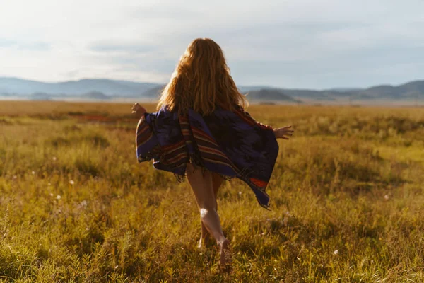 Vista desde atrás. Una chica pelirroja con ropa étnica corre por el campo bajo el sol del atardecer contra el telón de fondo de las montañas. — Foto de Stock