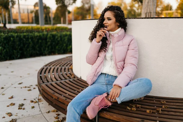 A menina brincalhão brooding senta-se no parque em um banco redondo e olha para o lado. Vestido com uma jaqueta rosa e botas rosa — Fotografia de Stock