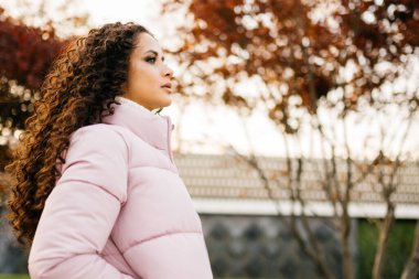 Sonbahar parkında kıpkırmızı yapraklı ağaçların arkasında uzun, kıvırcık saçlı, ceketli bir kız duruyor ve uzaklığa bakıyor.