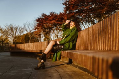 Kıvırcık saçlı şık bir güzellik kafasının üstünden dökülüyor sonbahar parkında bir bankta oturuyor kıpkırmızı ağaçların önünde.