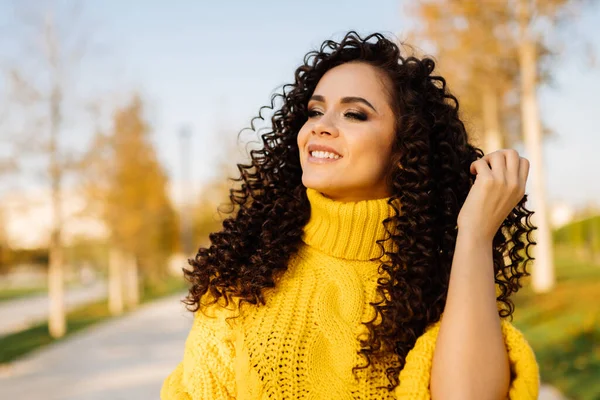 Stylově balí rukávy na tlustém žlutém svetru ručně pletené kudrnaté brunetky, usmívající se dívá do dálky v podzimním parku — Stock fotografie